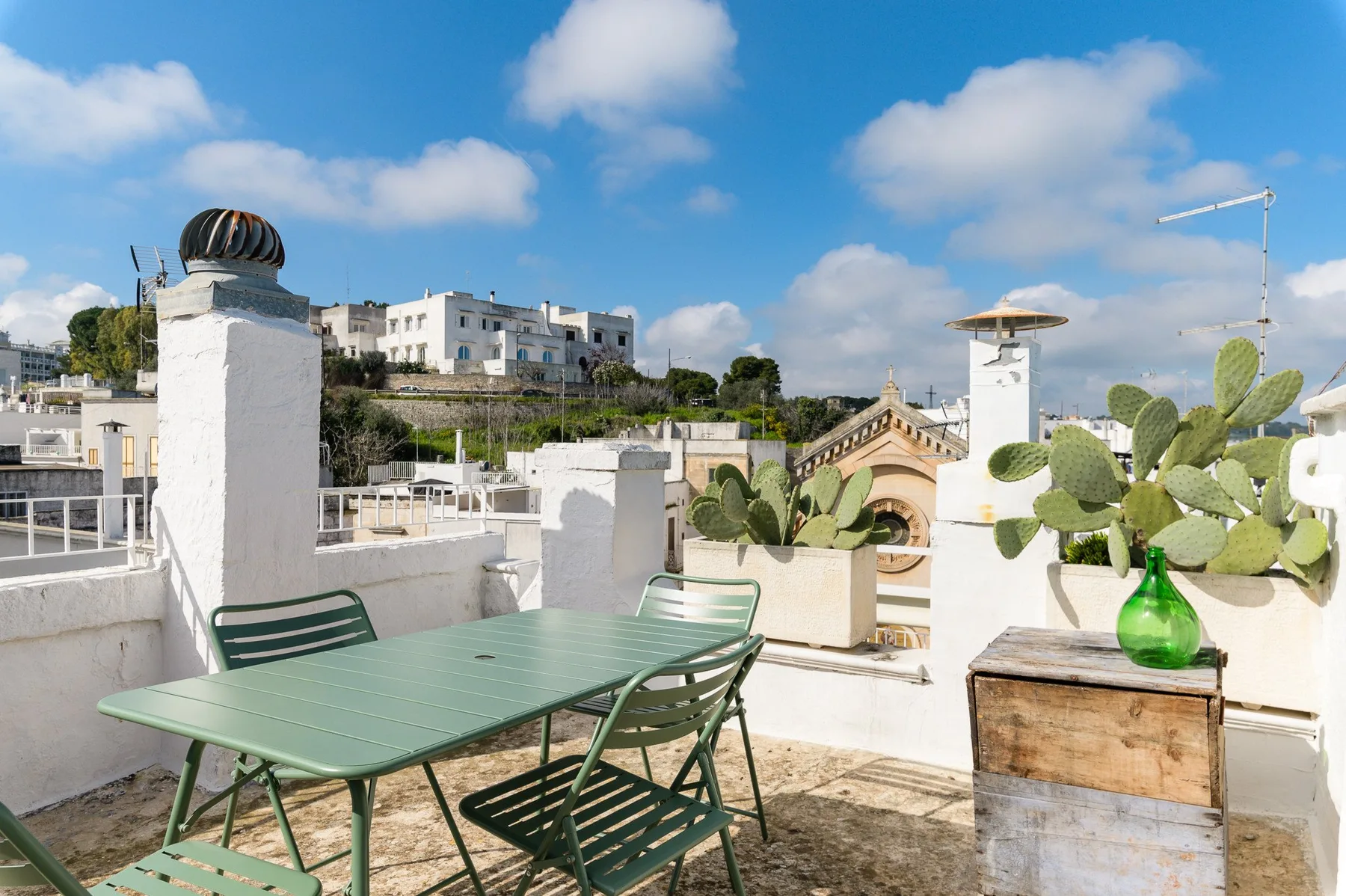 A sunny rooftop terrace with green seating, potted plants, and surrounding buildings.