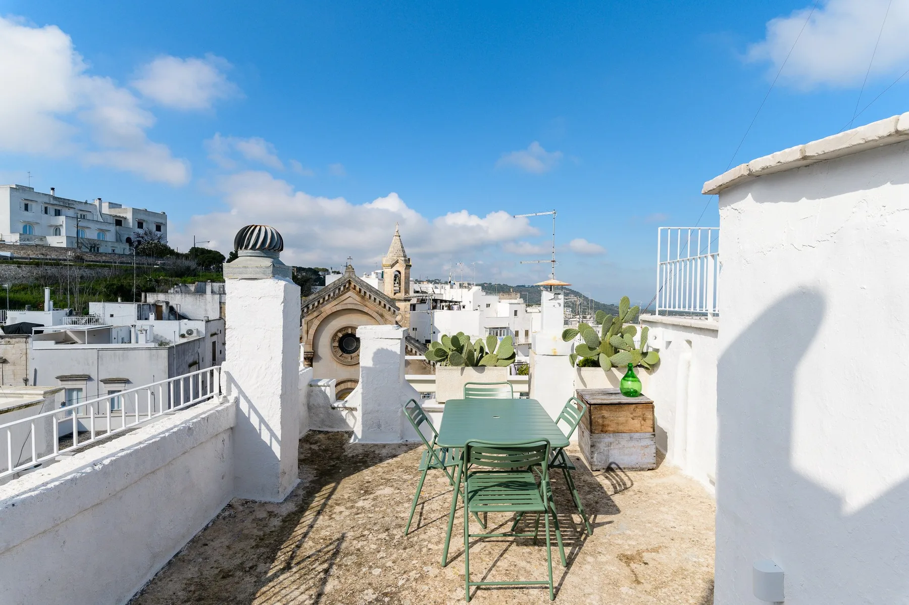 A rooftop terrace with green seating overlooks white buildings and a church tower.