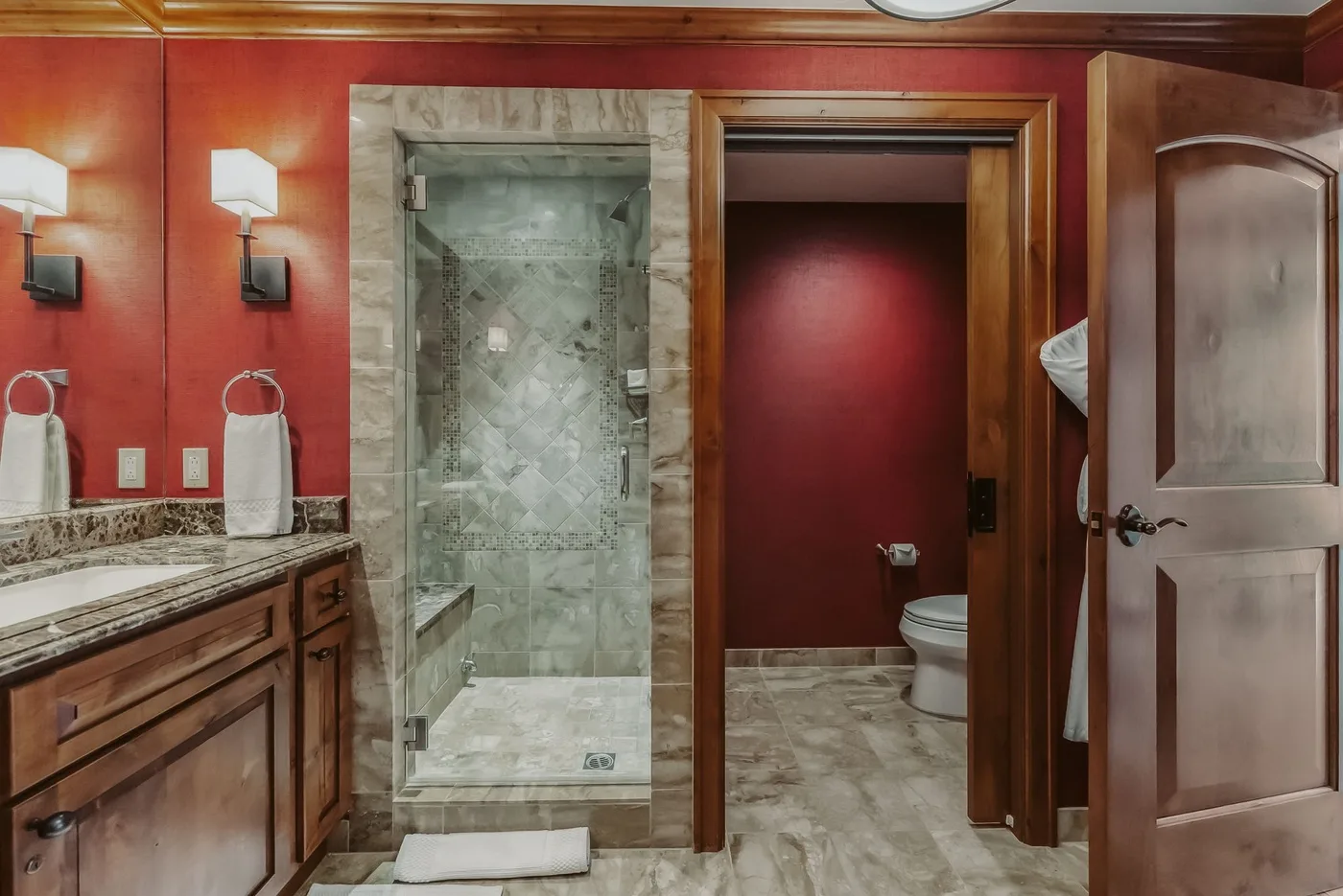 Un baño con ducha de mármol, tocador de madera y una puerta abierta hacia el área del inodoro.