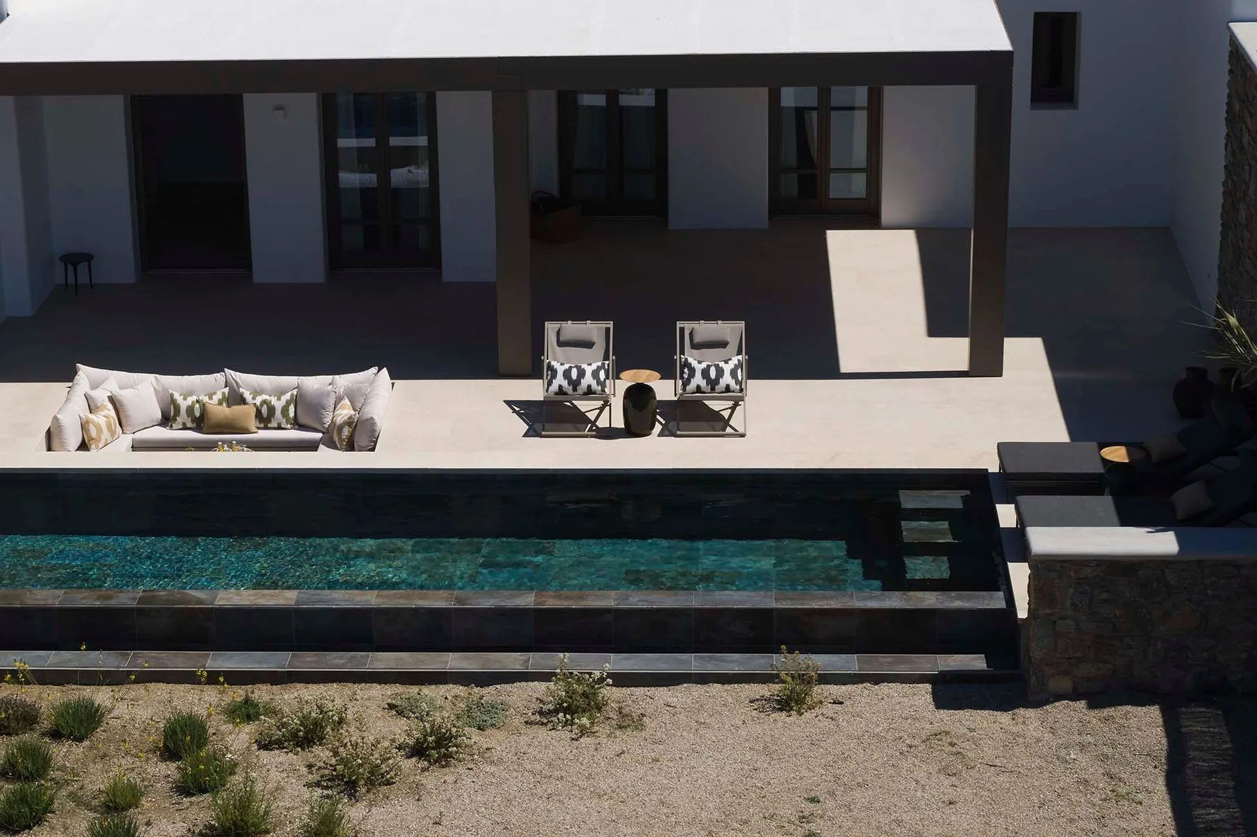 Un patio de villa moderna da a una piscina alargada, con asientos exteriores bajo un saliente sombreado.
