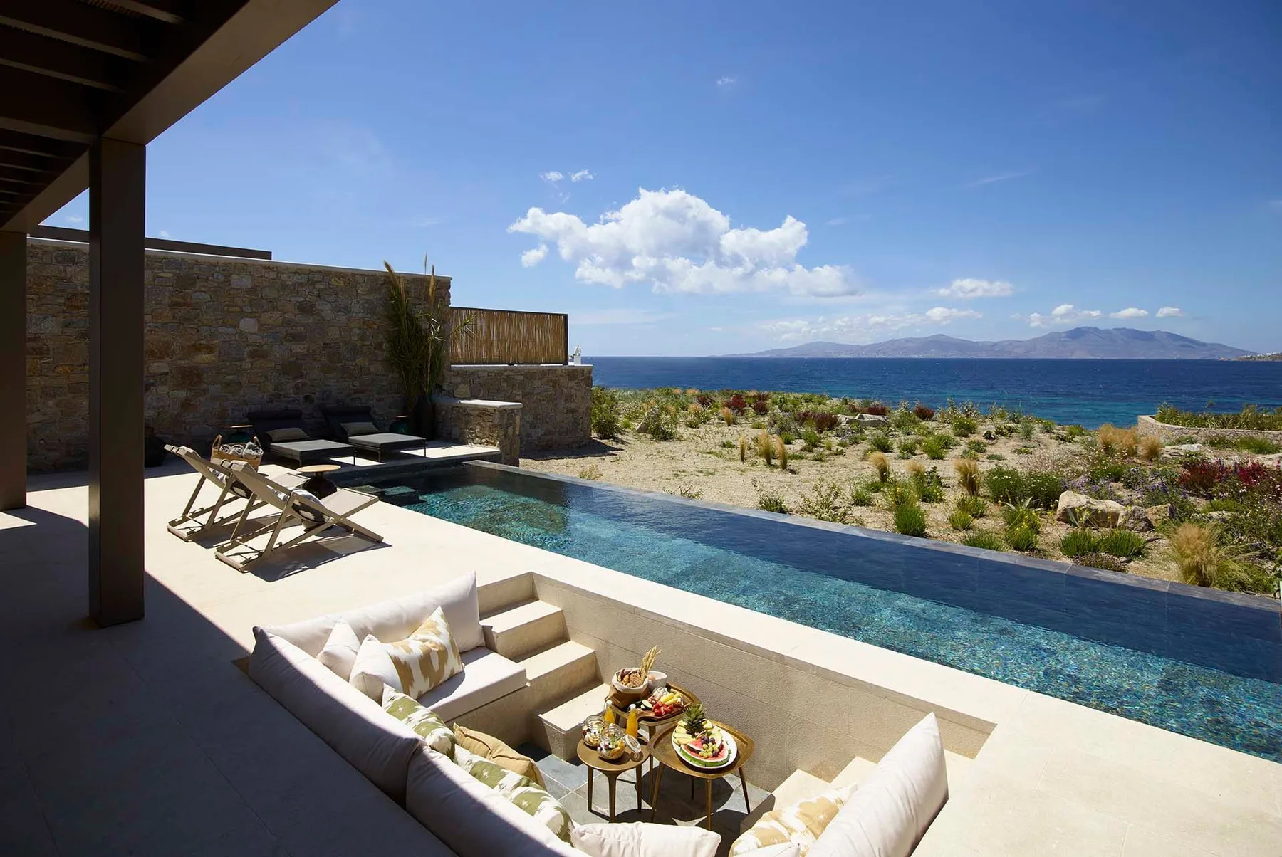 Una zona de estar exterior a la sombra da a una piscina estrecha y al mar.