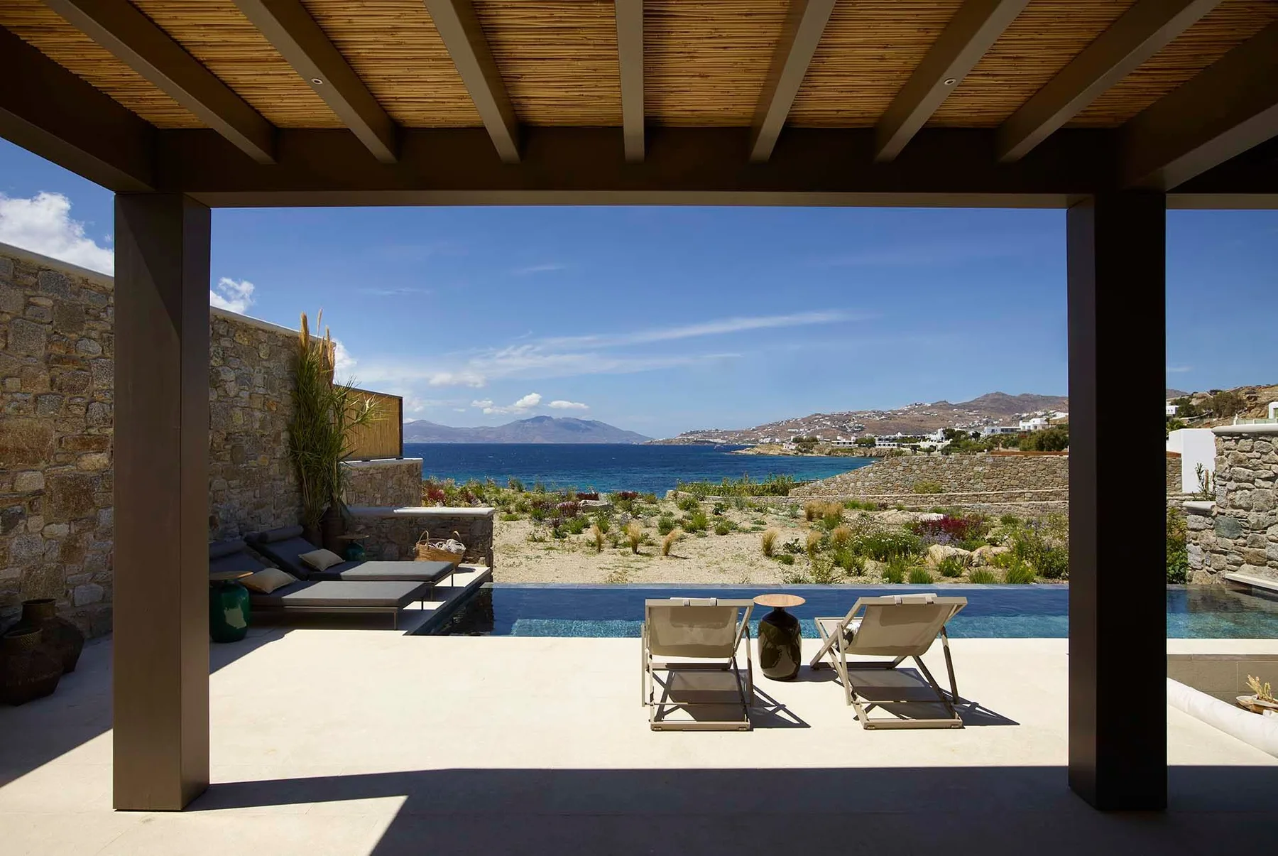 Una terraza sombreada da a una piscina, asientos y el mar.