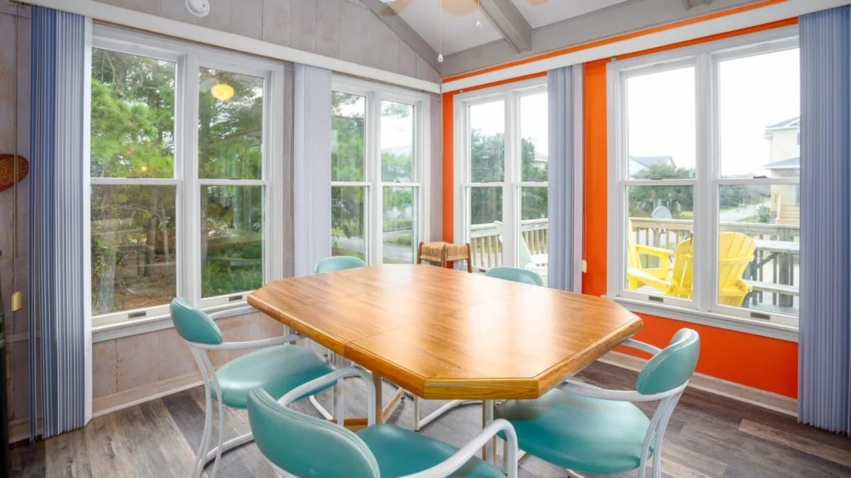 Un comedor luminoso tiene una mesa de madera y asientos turquesa junto a grandes ventanas.