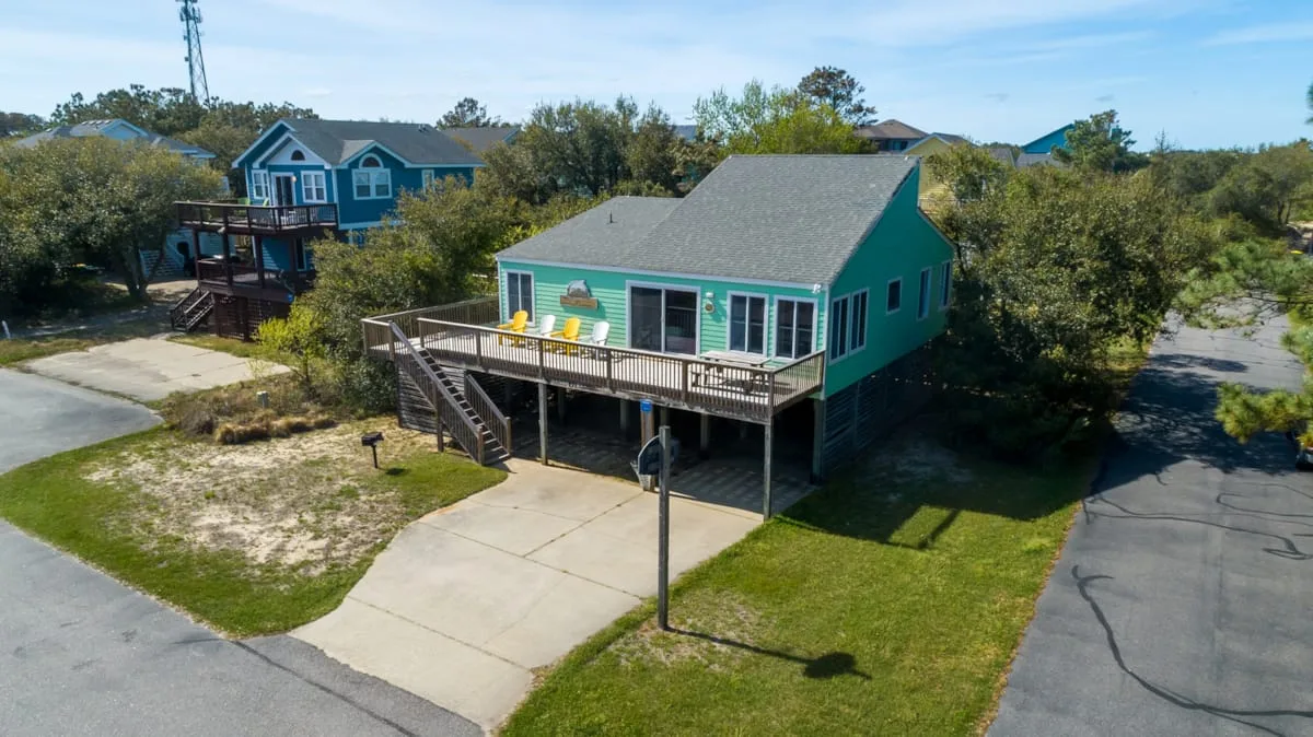 A teal house with a raised wooden deck sits beside a paved road.