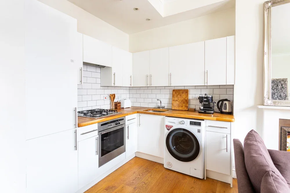A bright kitchen with white cabinets, a washing machine, and wooden flooring.