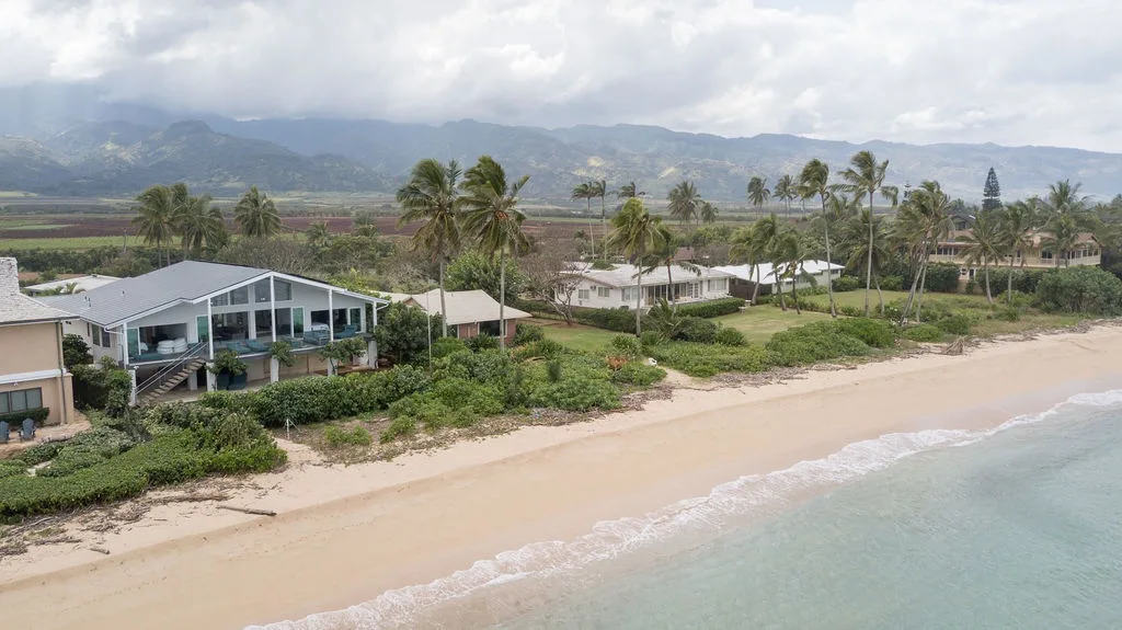 Una propiedad junto a la playa bordea una playa de arena con palmeras y montañas lejanas.