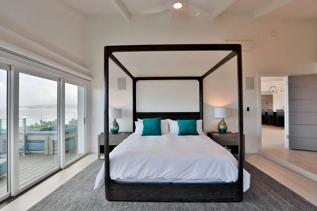 Un dormitorio luminoso tiene una cama junto a grandes ventanas con vistas al agua.