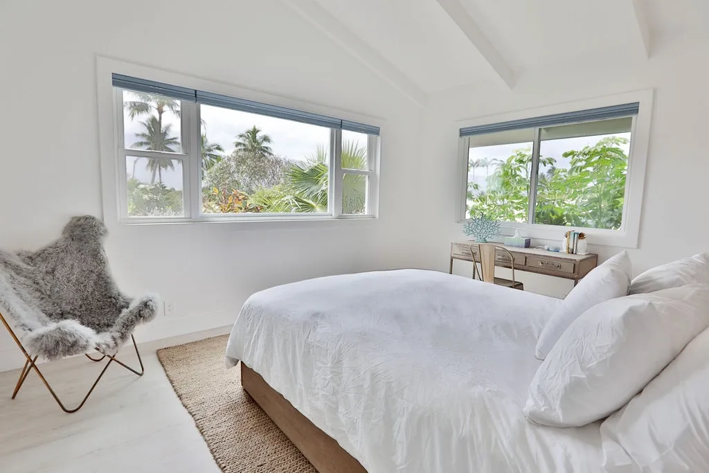 Un dormitorio luminoso con una cama, una silla y ventanas con vistas a vegetación frondosa.