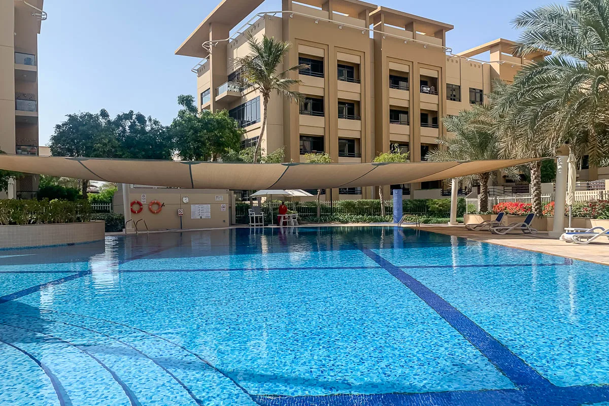 Una piscina al aire libre junto a un edificio de varios pisos, bajo toldos de sombra.