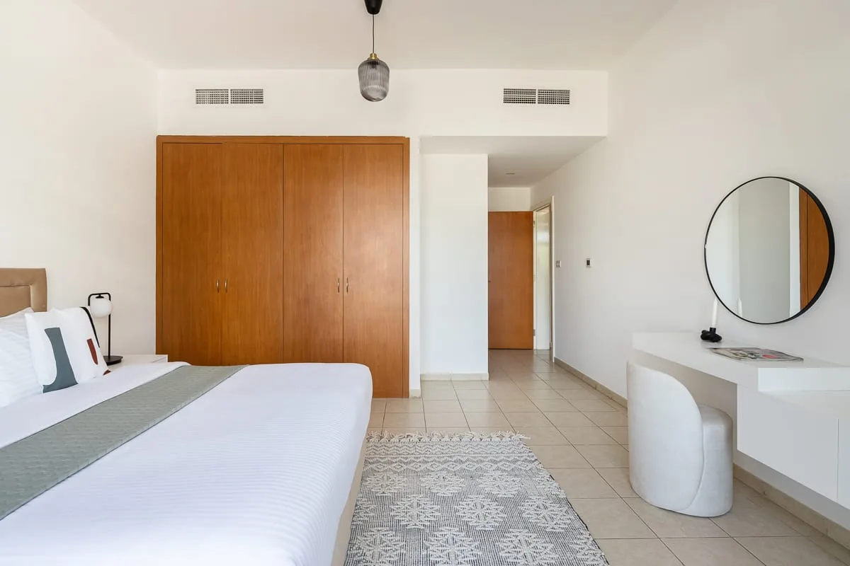 Un dormitorio luminoso con una cama, un armario de madera y un escritorio con espejo redondo.