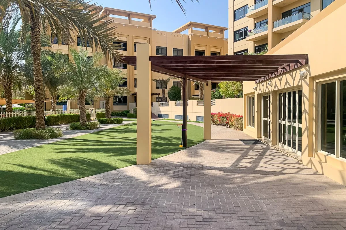 Un sendero pavimentado pasa bajo una pérgola junto a jardines y edificios de apartamentos.