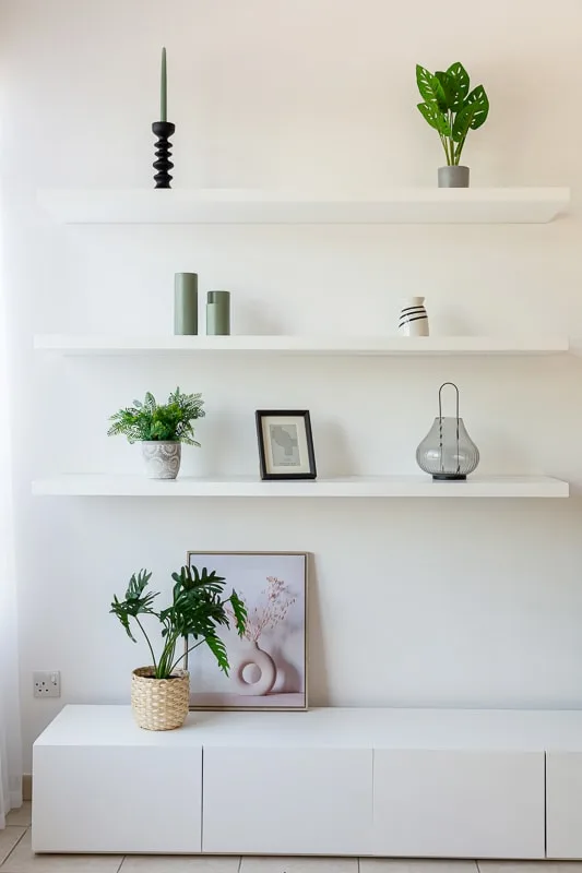 Una pared luminosa de sala tiene estantes blancos y un mueble bajo con plantas y adornos.