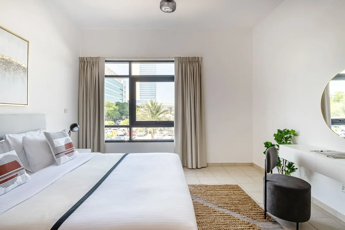 Un dormitorio luminoso con una cama grande junto a una ventana con vistas a árboles y edificios.