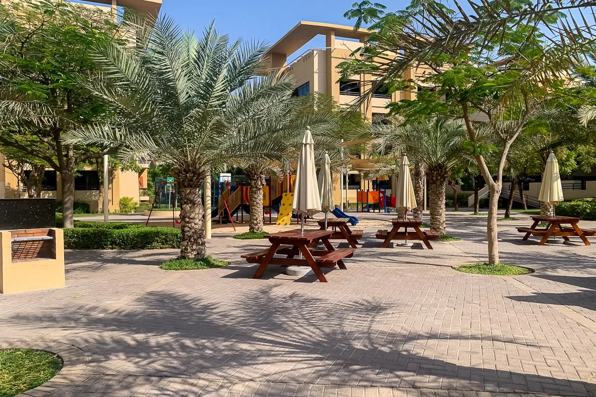 Un patio ajardinado al aire libre tiene mesas de picnic, palmeras y una zona de juegos infantil.