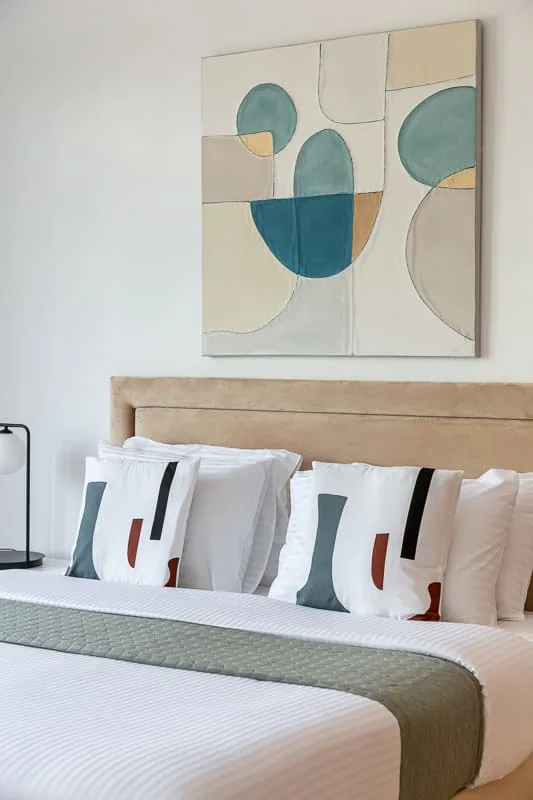 Una cama bien tendida con ropa de cama blanca y arte abstracto encima.
