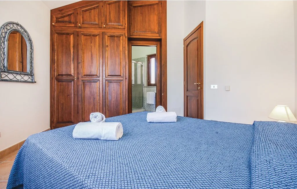 A bedroom with a blue-covered bed, wooden wardrobe, and doorway to a bathroom.