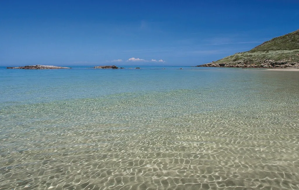 A shallow turquoise sea stretches toward low rocky islands and a green hillside.
