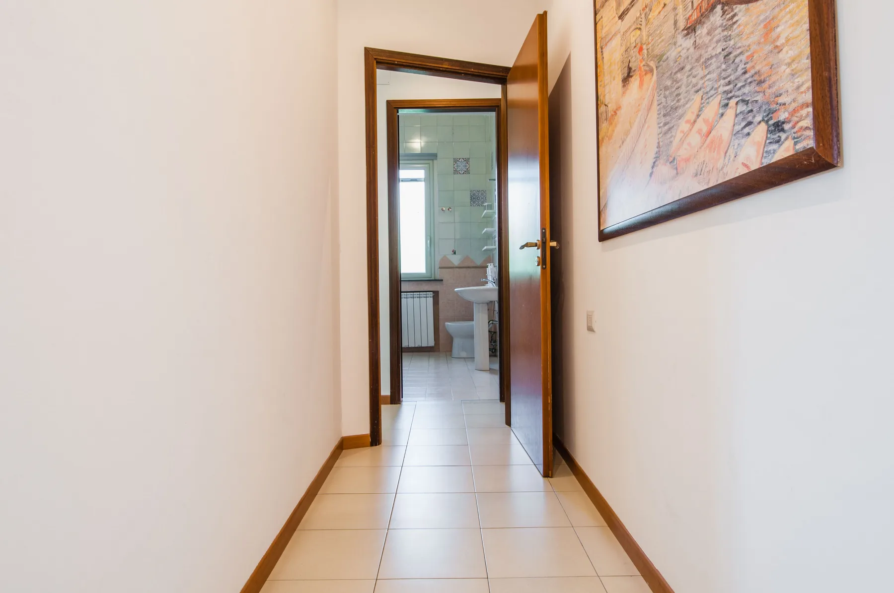 A tiled hallway leads to an open bathroom door, with a framed painting on the wall.