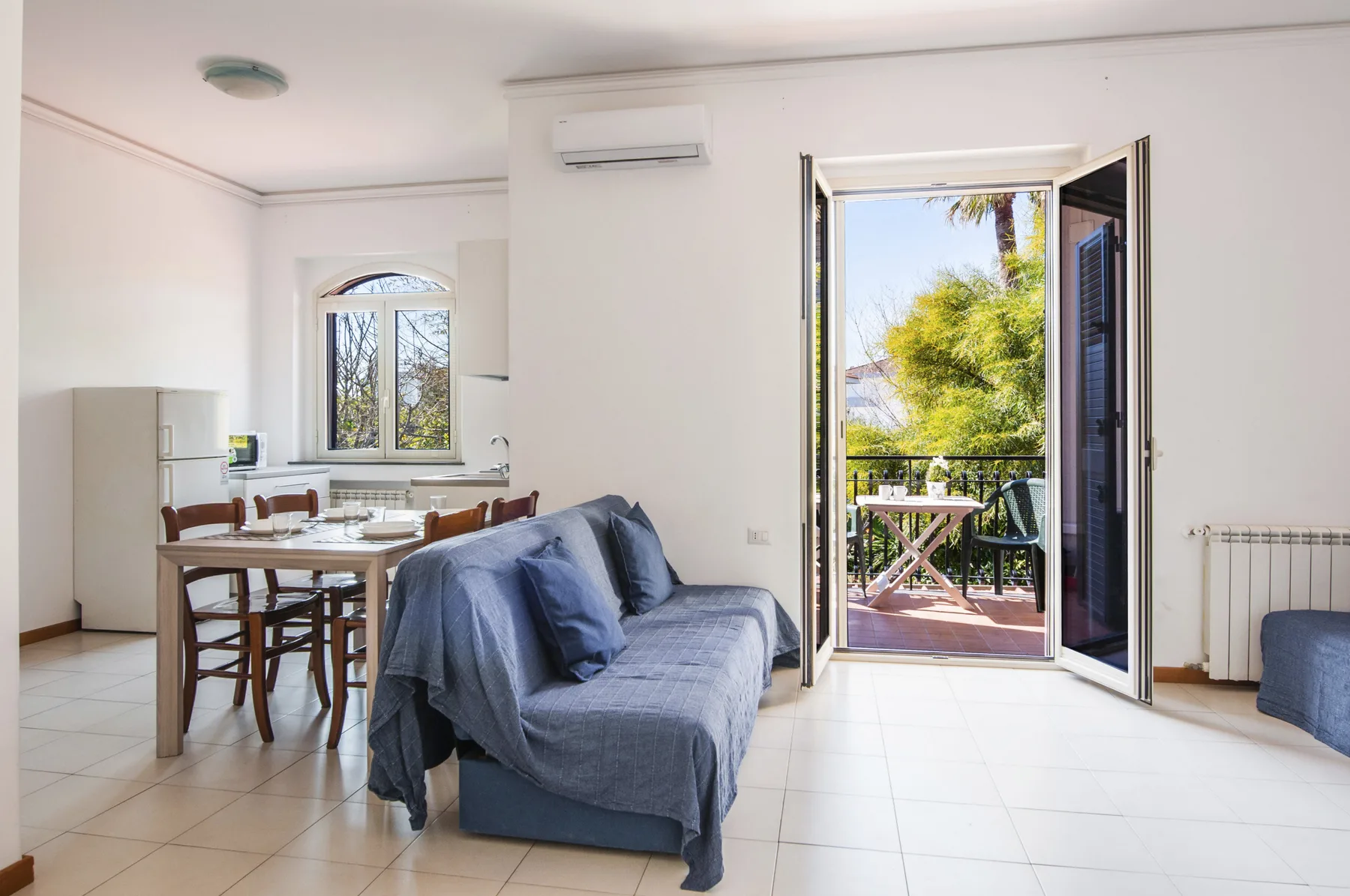 A bright living and dining area opens onto a balcony with greenery outside.
