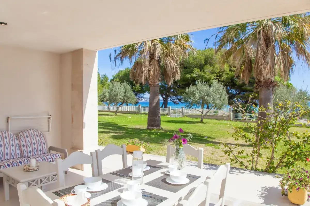 Una zona de comedor en un patio sombreado da a un jardín verde con palmeras.