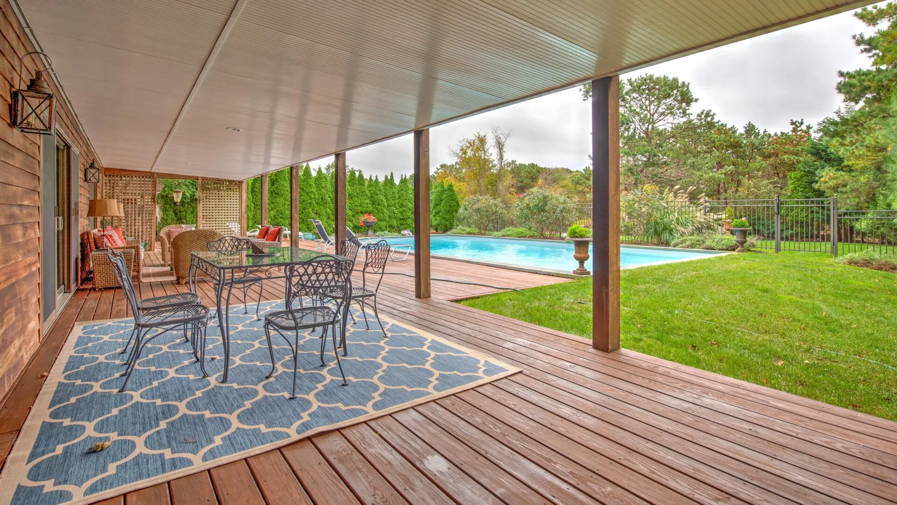 Un patio cubierto con asientos para comer da a una piscina y un jardín.