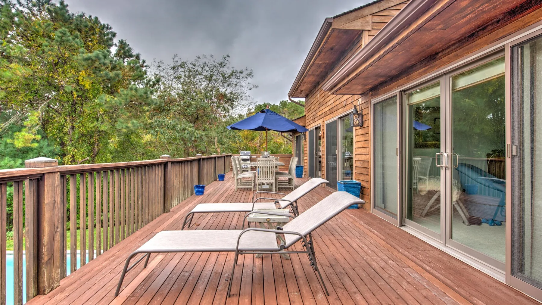 Una terraza de madera junto a la casa tiene asientos reclinables y un comedor bajo una sombrilla azul.