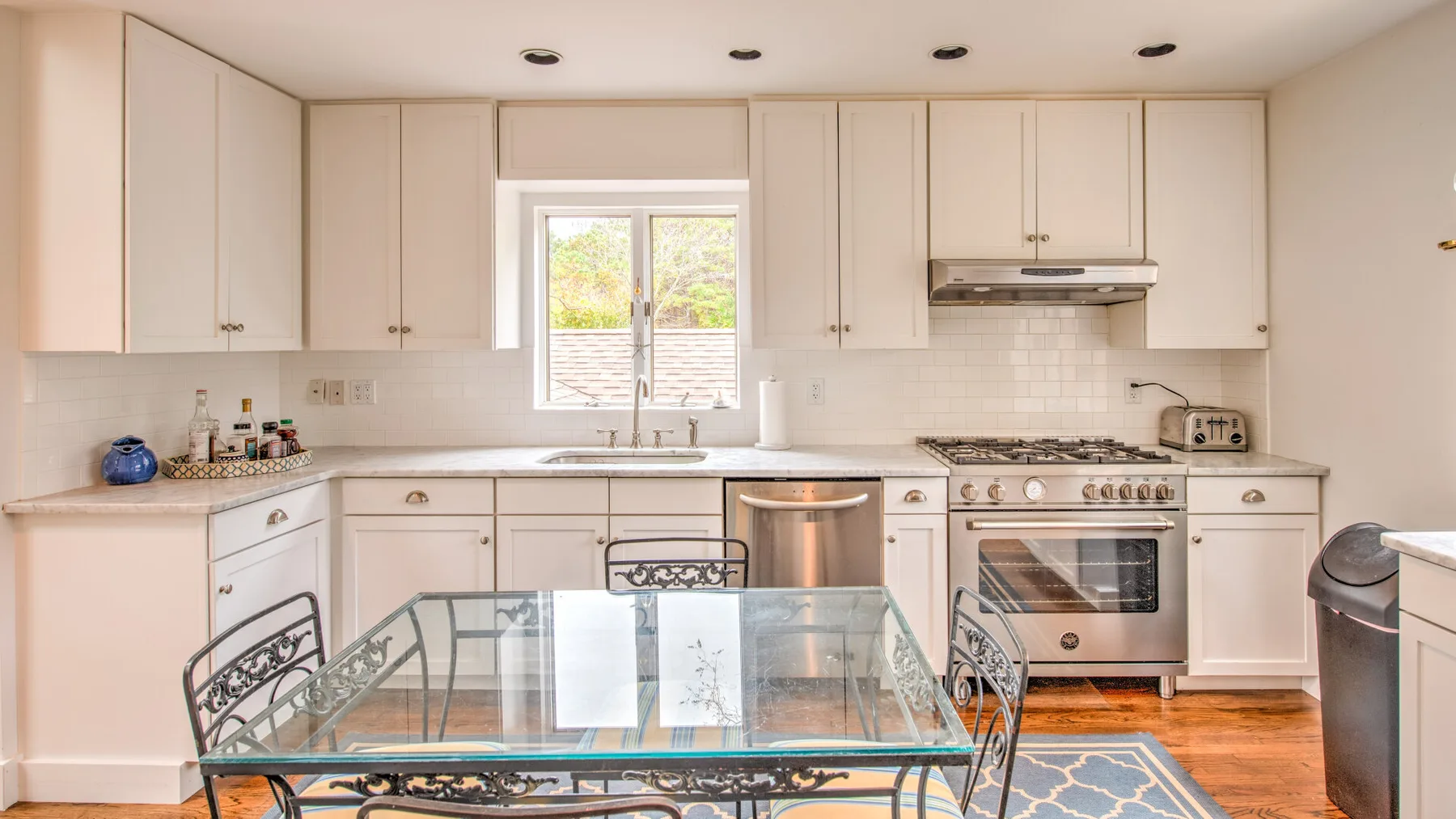Una cocina luminosa con gabinetes blancos, una ventana central y una mesa de comedor de vidrio.