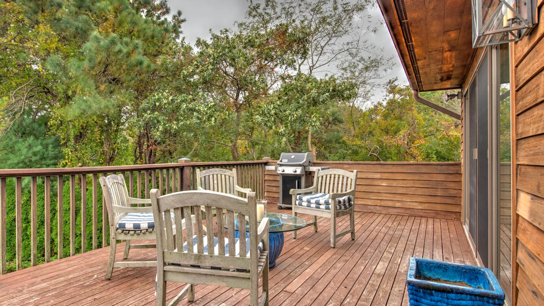 Una terraza de madera con asientos y una parrilla da a árboles frondosos.