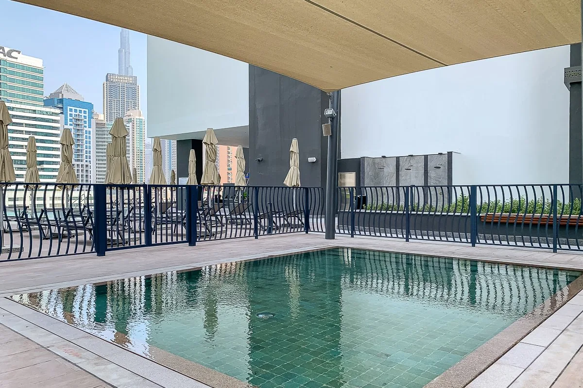 A rooftop swimming pool sits beside a railing with city buildings in the background.