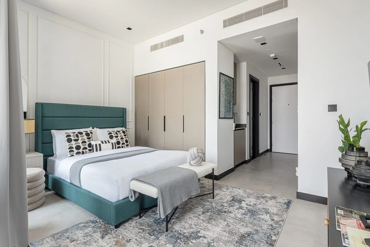 A bright bedroom with a teal bed, pale bedding, and a patterned rug.