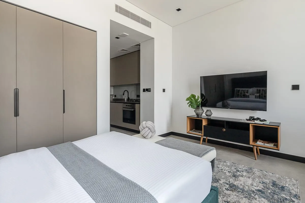 A bright bedroom with a neatly made bed, television, and compact kitchenette.
