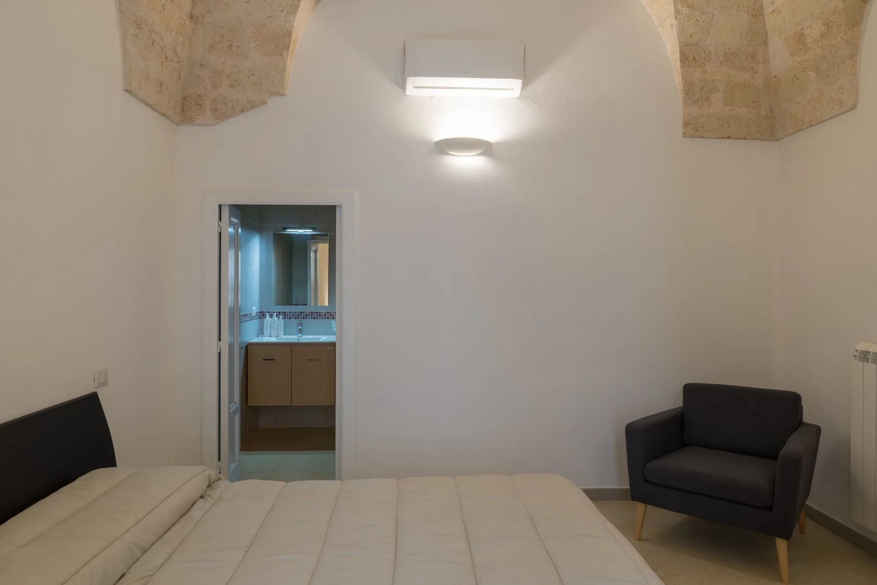 A bedroom with a bed and an armchair, with a bathroom visible through the doorway.