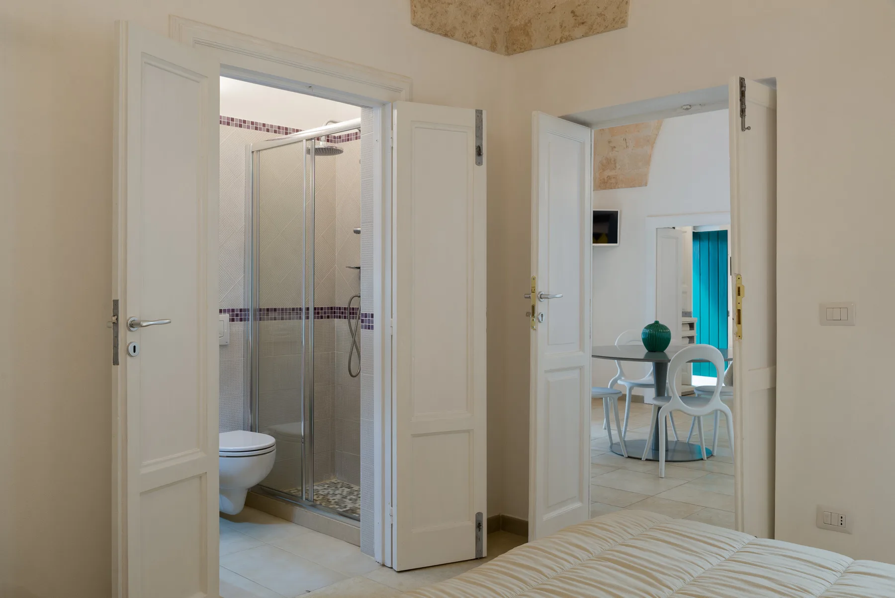 A bedroom with open doors overlooking a bathroom and a dining area.