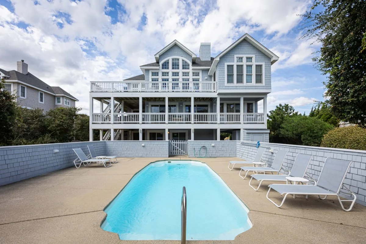 A backyard swimming pool sits in front of a large house with raised decks.
