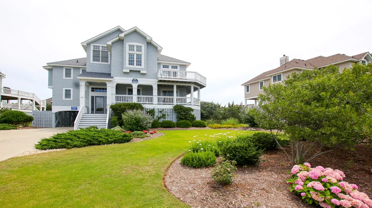 A large blue house stands behind a landscaped lawn and flowering shrubs.