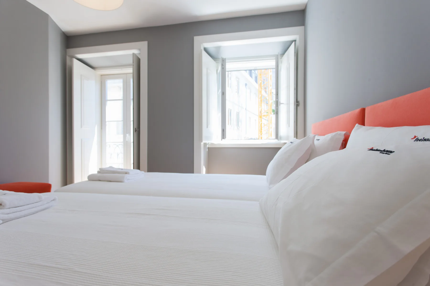 Un dormitorio luminoso con ropa de cama blanca y ventanas con vistas a edificios cercanos.
