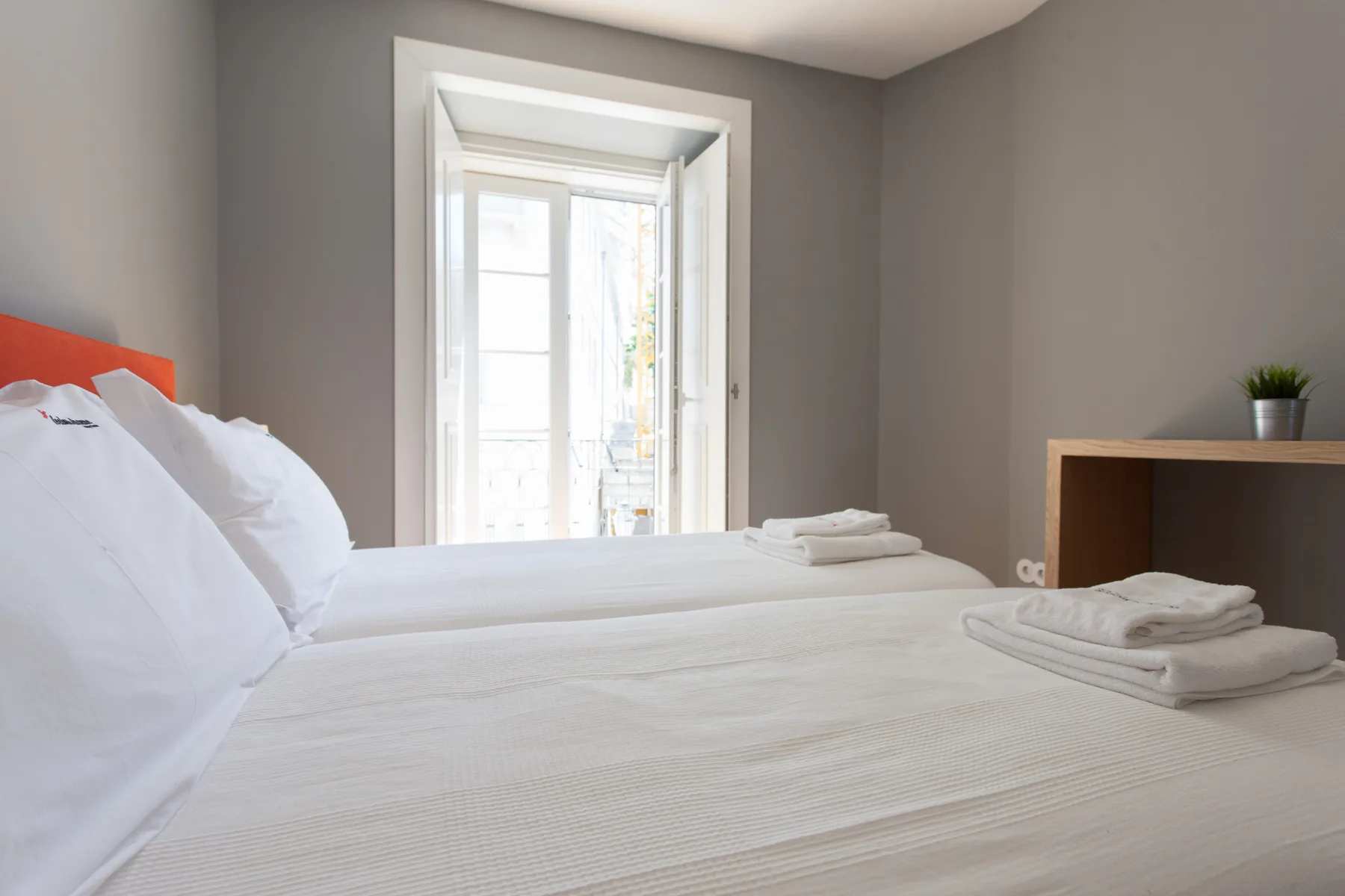 Dormitorio con ropa de cama blanca, una ventana luminosa y una pequeña repisa de madera.