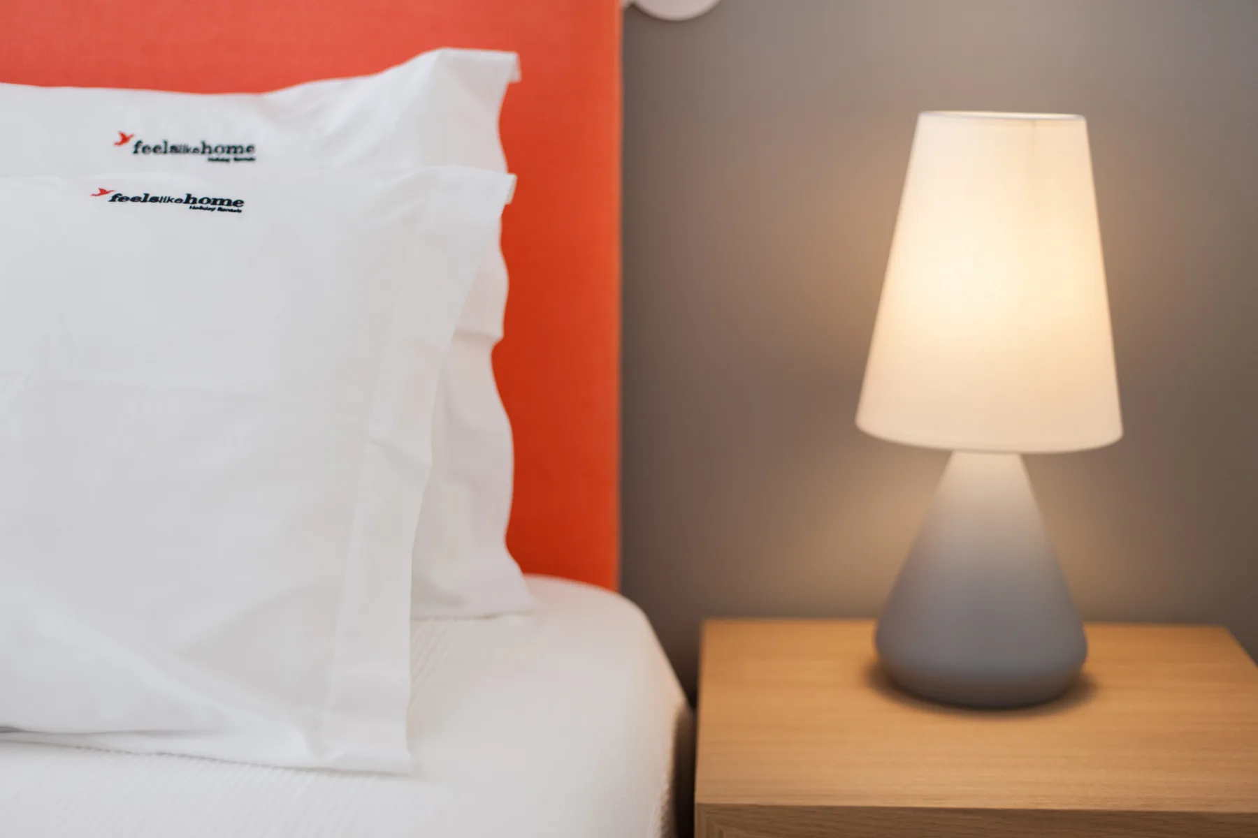Una cama con almohadas blancas está junto a una lámpara de mesa encendida.