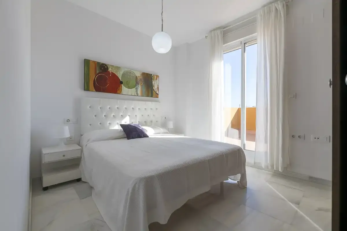 A bright bedroom with a neatly made bed and a glass door to the balcony.