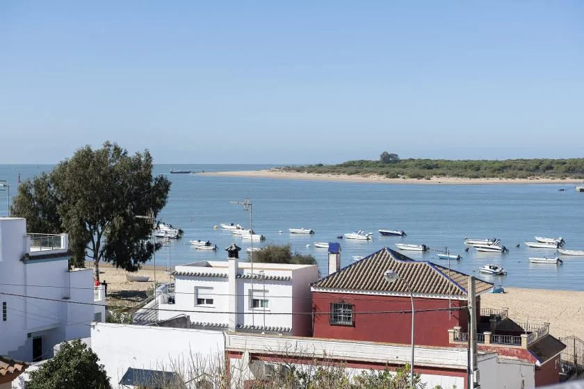 Coastal waterfront with low buildings, a sandy shore, and boats on calm water.