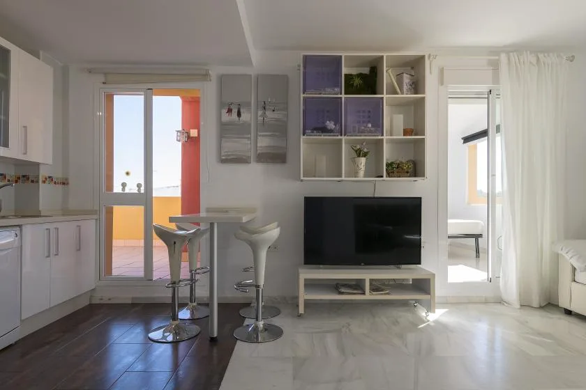 A bright open-plan living area with a television, breakfast bar, and kitchen.