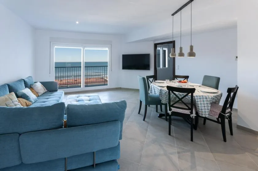 A bright living and dining area has blue seating, a dining table, and balcony doors overlooking the sea.