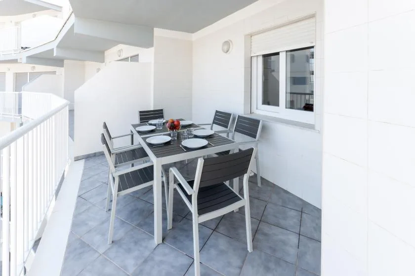 A tiled balcony has an outdoor dining table and seating beside a window.