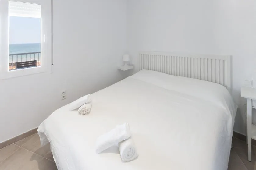 A bright bedroom with a white bed and a window overlooking the sea.