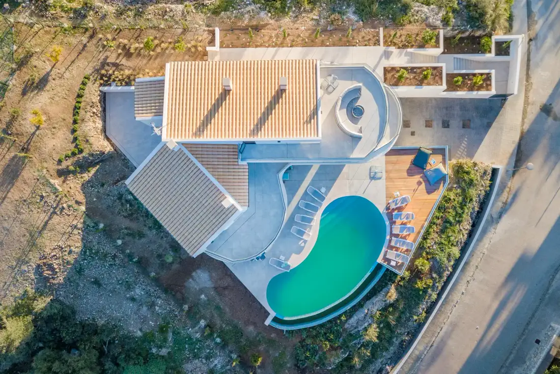 An aerial view shows a villa with a curved swimming pool and a paved terrace.