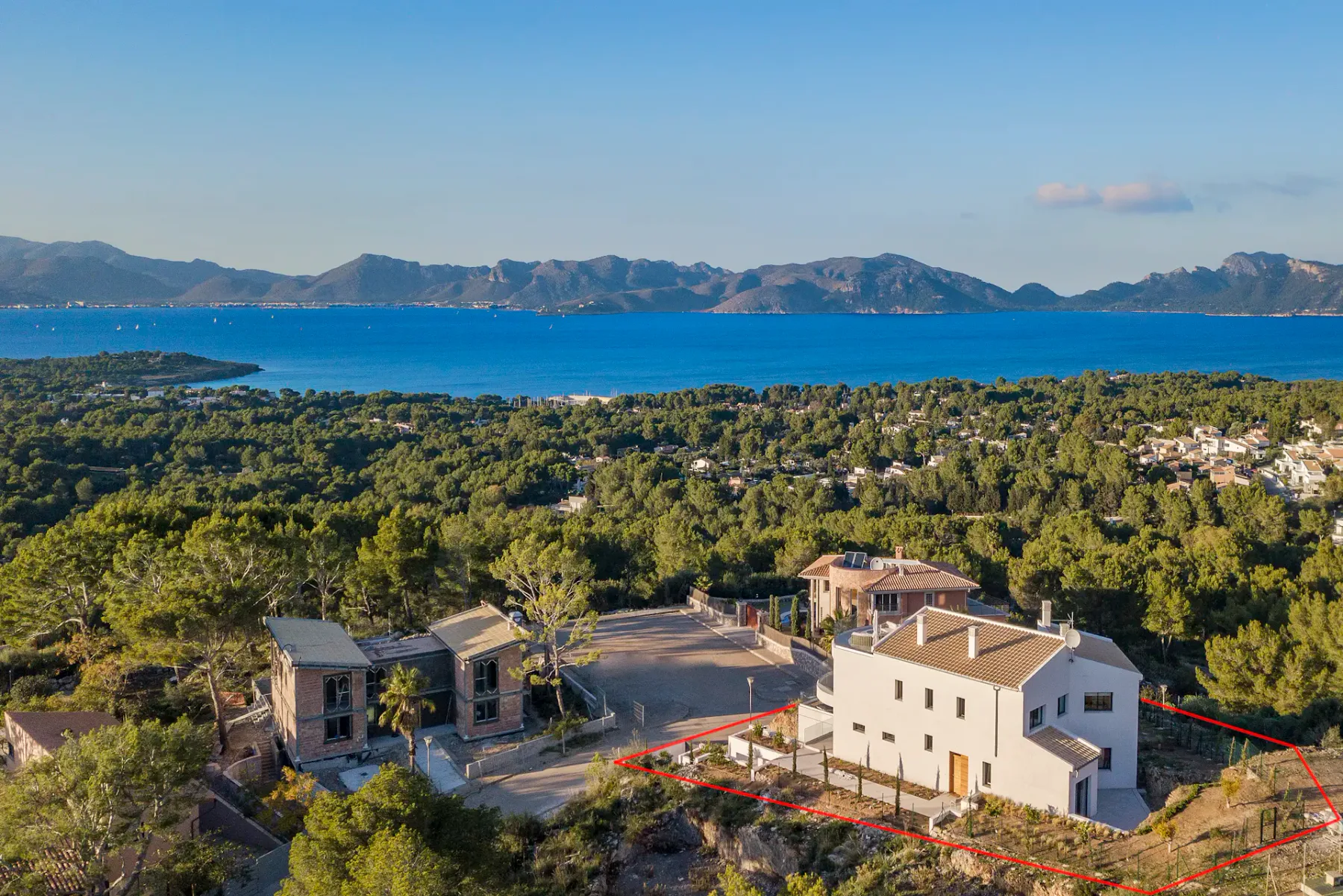 An aerial view shows a villa beside wooded hills, with the sea and distant mountains beyond.