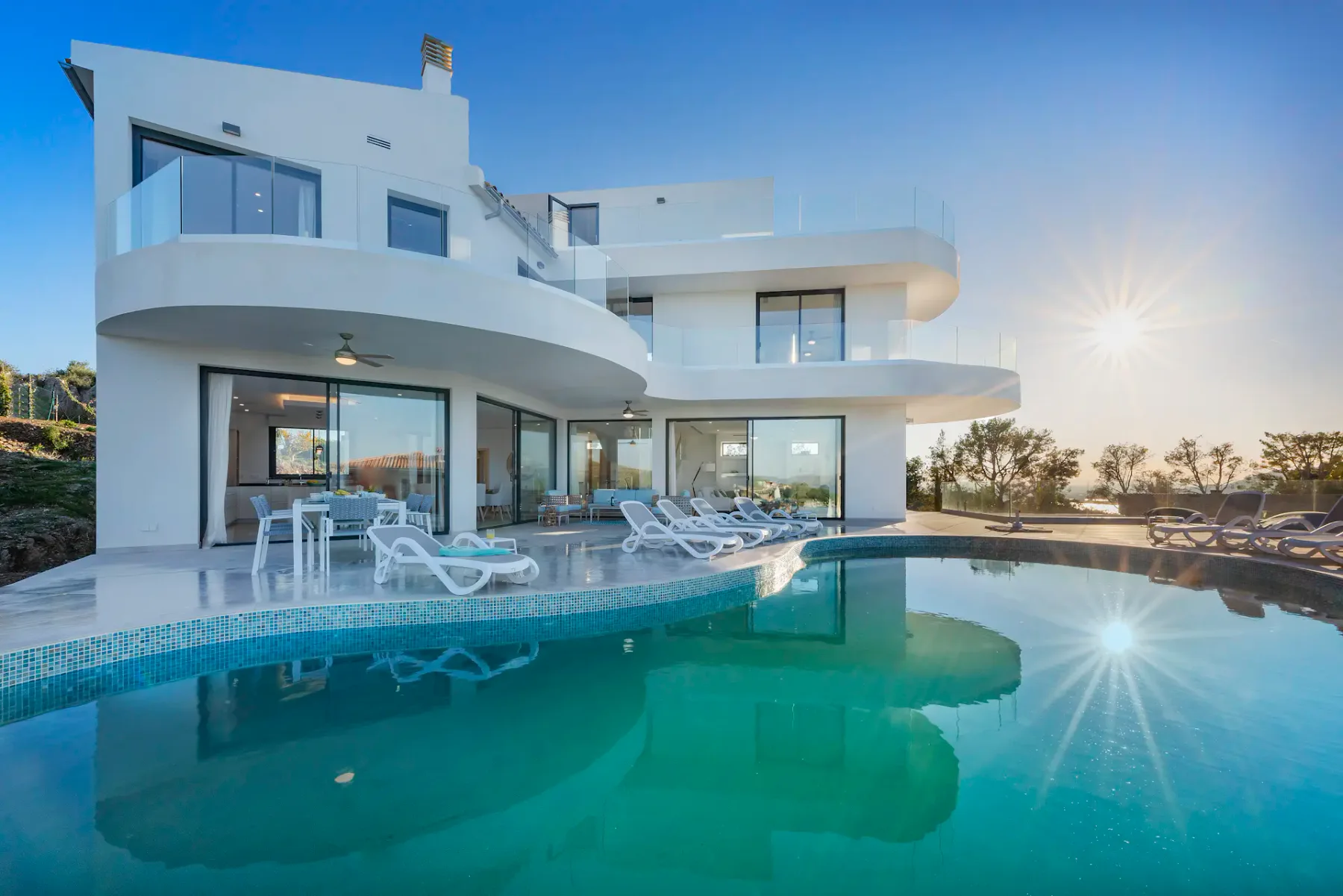 A modern white villa overlooks a swimming pool with lounge seating beside it.