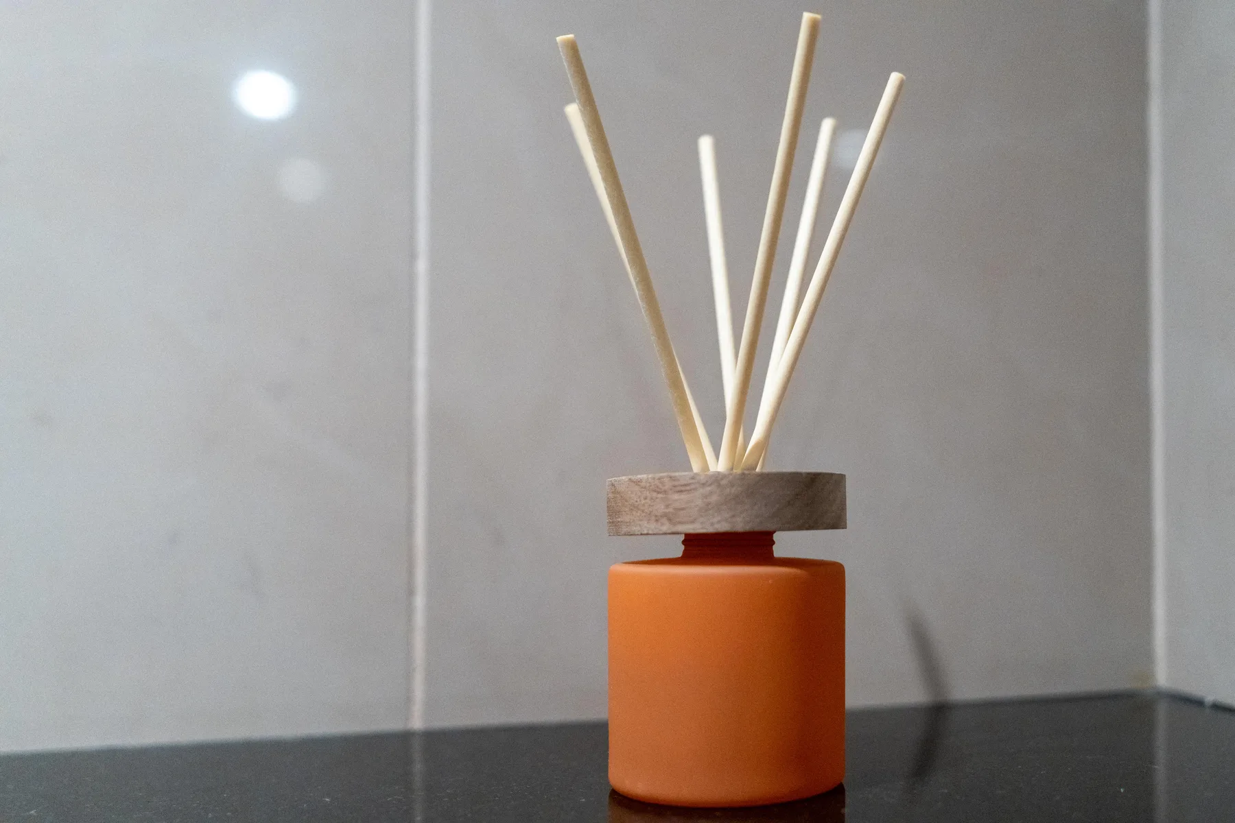 Un difusor de varillas naranja está sobre una encimera oscura frente a una pared clara.