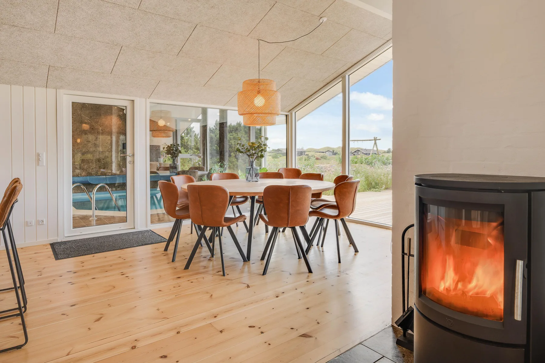 Comedor luminoso con una mesa de madera, asientos y una estufa encendida junto a grandes ventanas.
