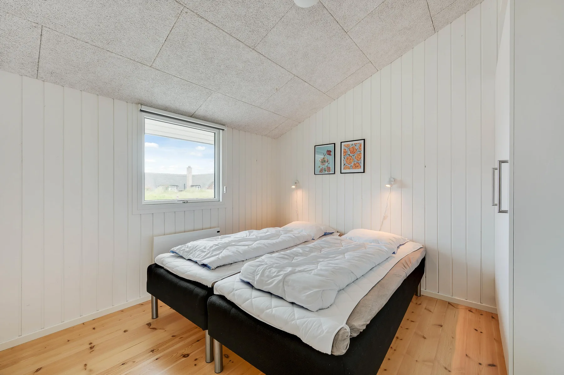 Un dormitorio luminoso con una cama bajo una ventana y cuadros en la pared.